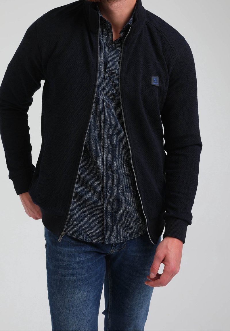 Giacca zip con finitura tessurizzata di colore blu navy; indossata sopra una camicia blu con motivo. Vestibilità slim, polsini a coste e dettaglio del logo sul petto.