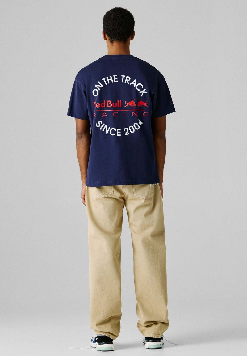 Άνδρας με μπλε μπλουζάκι με το κείμενο "On the Track Red Bull Racing Since 2004" στην πλάτη, μπεζ παντελόνι και μαύρα αθλητικά παπούτσια με λευκές σόλες.