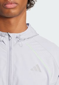 Lichtgrijze zip-up jas met een capuchon, mesh-gepatroneerd zijpaneel, reflecterende accenten en subtiele branding op de borst.