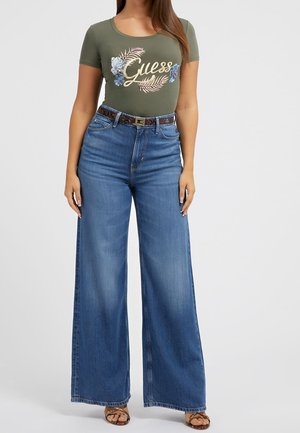 Green t-shirt with floral "Guess" logo, paired with high-waisted, wide-leg blue jeans and a brown patterned belt. Textured sandals complete the look.