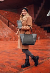 Black tote bag with textured accents, paired with a gray phone and gloves. The model wears a tan coat and black lace-up boots. Brick background.
