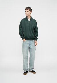 Zelená mikina se čtvrtzipem, kombinovaná se světle modrými volnými džínami a černými teniskami s bílými akcenty. Jednoduchý, neformální design.