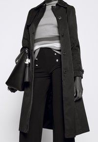 Abrigo negro de trench con botones, superpuesto a un suéter de cuello alto a rayas gris y blanco. Pantalones negros de tiro alto con botones decorativos. Bolso negro.
