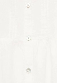 Tela blanca con tres botones de nácar alineados verticalmente, mostrando detalles de costura en un panel de prenda texturizado.
