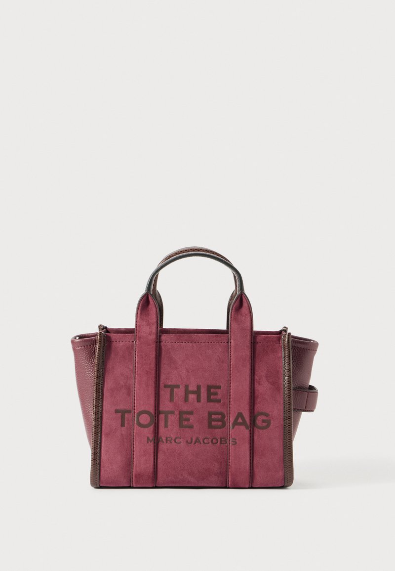 Bordeaux ruskind- og lædertote med korte håndtag og forrest tekst, der læser "THE TOTE BAG MARC JACOBS" på en hvid baggrund.