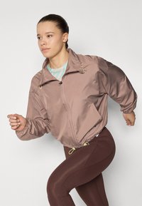 Giacca cropped rosa chiaro con polsini elasticizzati, zip frontale e cordini regolabili. Abbinata a leggings marrone scuro, con un design sportivo.