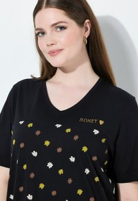 Žena se rovnými hnědými vlasy a zlatými kruhovými náušnicemi, oblečená v černém tričku ve tvaru V s malými žlutými, bílými a hnědými květinami a nápisem "HONEY".