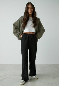 Přesně vyhovující olivově zelená bomber bunda přes bílý crop top, spárovaná s černými kalhotami volného střihu a stříbrnými teniskami. Hladké textury, ležérní styl.