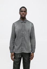 Tânăr stând cu fața în față, purtând o cămașă gri decolorată cu nasturi și pantaloni negri cu buzunare mari în față.