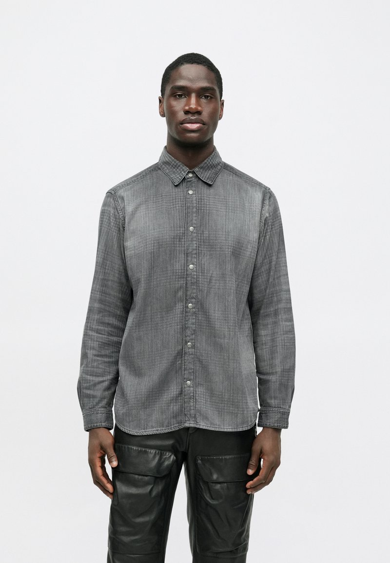 Tânăr stând cu fața în față, purtând o cămașă gri decolorată cu nasturi și pantaloni negri cu buzunare mari în față.