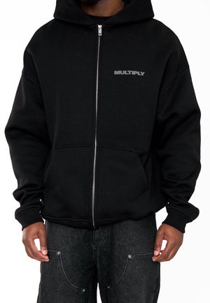 Felpa nera con zip e logo "MULTIPLY" sul petto, indossata con pantaloni di jeans scuri, mostrata dalla nuca fino a metà coscia su una persona in piedi.