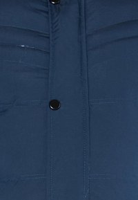 Tamsiai mėlyna pašiltinta striukė su horizontaliomis siūlėmis, juodais mygtukais ir lygiu paviršiumi, su šoninėmis kišenėmis.