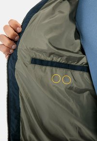 Ręka trzymająca otwartą granatową kurtkę ukazującą gładką oliwkową podszewkę z wewnętrzną kieszenią oraz małymi drukowanymi ikonami wodoodporności i odporności na plamy.