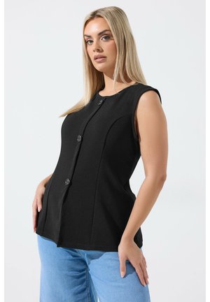 Mujer rubia con top negro sin mangas abotonado y vaqueros azul claro, posando con las manos cerca de las caderas frente a un fondo liso.