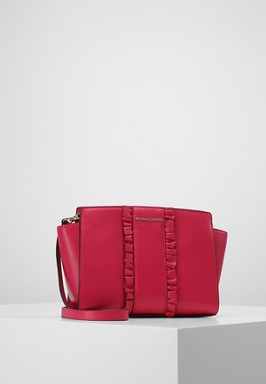 Czerwona torebka Michael Kors z pionowym marszczonym paskiem i odpinanym paskiem na ramię, prezentowana na białym piedestale na szarym tle.