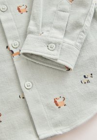 Camiseta gris claro hecha de tela suave, con un patrón de pequeñas ilustraciones coloridas de perros y botones blancos.