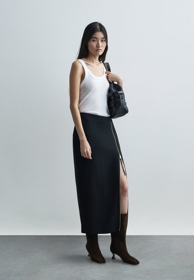 Nő fehér ujjatlan felsőben, fekete szoknyában, melynek magas oldalsó cipzárhasítéka van, barna magassarkú csizmával, és fekete táskát tart a vállán.