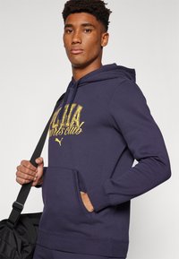 Marineblå hættetrøje med kangaroo-lomme, prydet med gul grafisk tekst "Sports Club" og et logo. Blød stof, afslappet pasform.