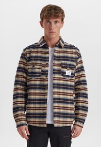 Camicia di flanella a quadri blu, marrone e crema. Presenta due tasche sul petto, chiusura con bottoni e un'etichetta sulla tasca sinistra. Tessuto testurizzato.