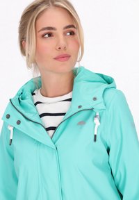 Egy türkiz vízálló dzseki kapucnival és ezüst színű szerelvényekkel. Elöl cipzárral, zsinórokkal rendelkezik, és egy esernyő logó látható a mellkason.