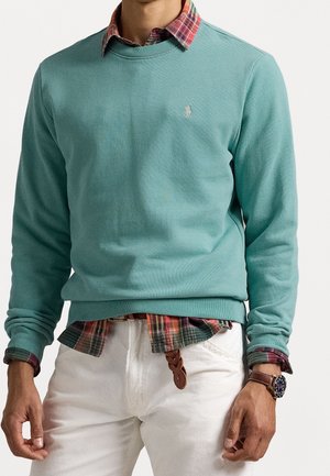 Man wearing teal crewneck sweater over red plaid shirt with white pants and brown braided belt, showing wristwatch on left hand.