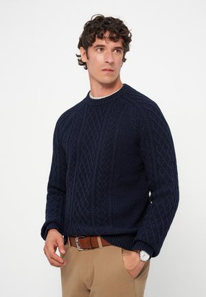 Jeune homme aux cheveux bouclés porte un pull torsadé bleu marine, un pantalon beige, une ceinture marron et une montre-bracelet, posant sur un fond blanc.
