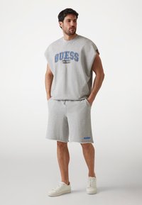 Felpa grigia in cotone con logo blu "GUESS", abbinata a pantaloni corto grigi coordinati. Texture morbida, vestibilità sportiva e sneakers bianche completano l'outfit.