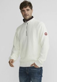Witte, gestructureerde, ritssluiting trui met een hoge kraag en geribde manchetten. Heeft een klein rood logopatch op de linker mouw. Draagt met blauwe jeans.