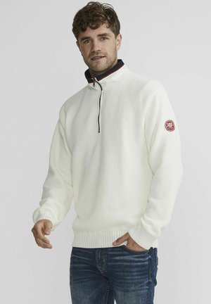 Hvid, struktureret, lynlåset sweater med høj krave og ribbede manchetter. Har et lille rødt logo-patch på venstre ærme. Båret med blå jeans.