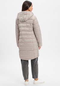 Gevoerde lichtbeige jas met capuchon, verticale streepdetails en elastische manchetten. Draagt over grijze broek en witte sneakers.