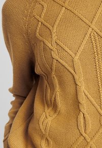 Pull en tricot jaune moutarde avec un motif texturé de câbles entrelacés et un ourlet côtelé. Présente un tissu en tricot grossier et épais.