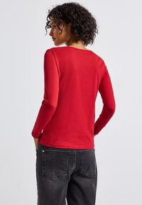 Frau mit lockigem Haar, die ein figurbetontes rotes Langarmoberteil und schwarze Jeans trägt, mit dem Rücken zu einer einfarbigen hellen Hintergrundwand gewandt.