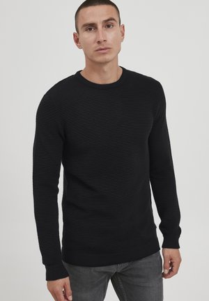 Black ribbed sweater with long sleeves and a crew neckline, featuring a textured pattern and fitted cuffs. Worn with gray jeans.