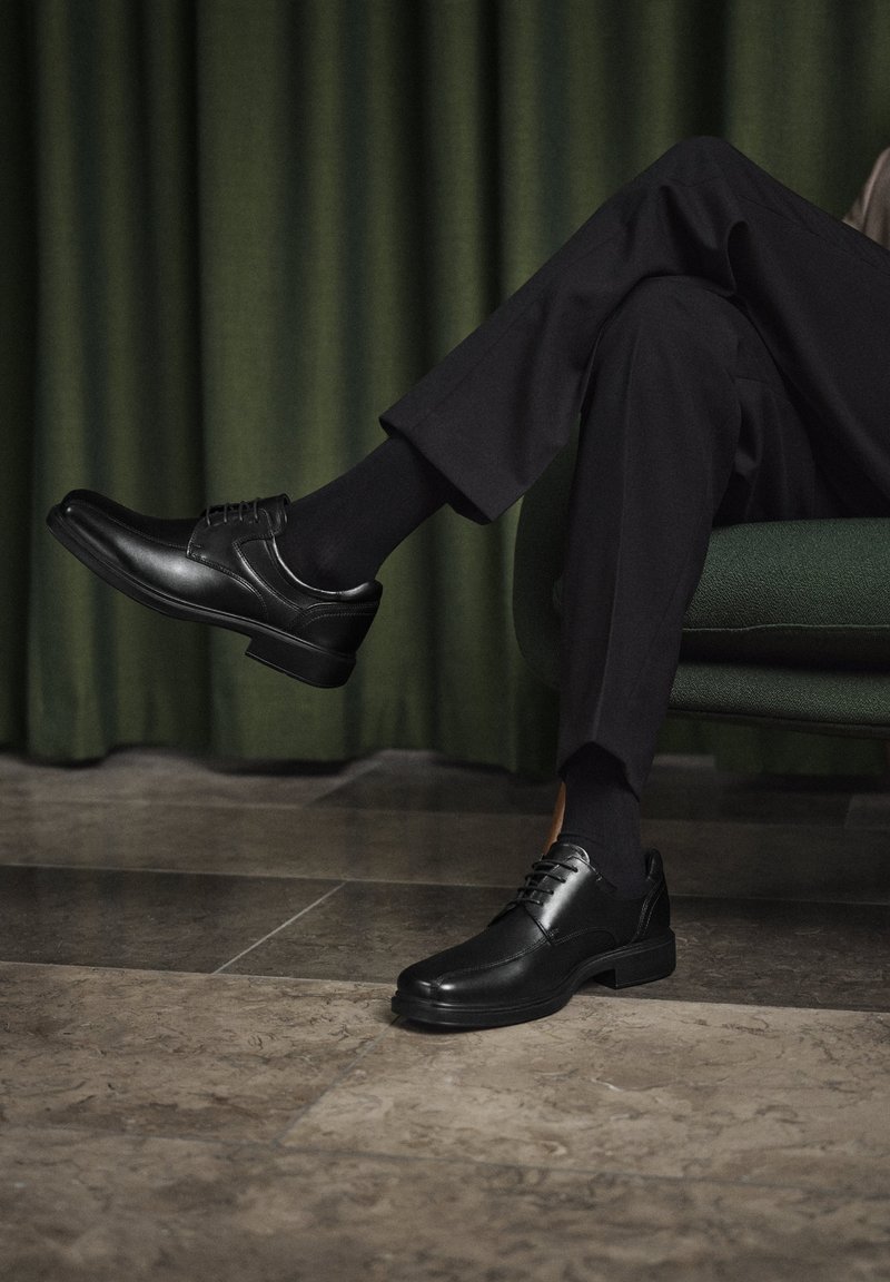 Personne portant des chaussures habillées noires, des chaussettes noires et un pantalon foncé, assise jambes croisées sur une chaise tapissée verte devant des rideaux verts.