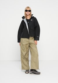 Črna kratka jakna z sivo podlogo, zadrgo in dvema sprednjima žepoma, v kombinaciji z ohlapnimi bež cargo hlačami in sivimi pohodniškimi čevlji.