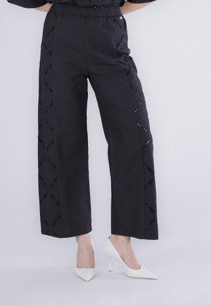 Pantaloni neri a gamba larga con motivi a forma di diamante sui lati, indossati con tacchi bianchi a punta.
