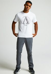 Valkoinen lyhythihainen T-paita, jossa on pyöreä musta ja vaaleanpunainen graafinen muotoilu, yhdistettynä harmaisiin housuihin ja mustiin urheilukenkään.