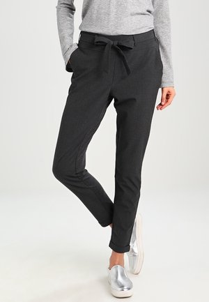 Persoon draagt nauwsluitende donkergrijze broek met een strikceintuur in de taille, omgeslagen zoom, grijze longsleeve en metallic zilveren instapschoenen.