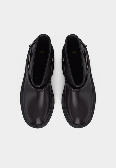 Ζευγάρι μαύρες δερμάτινες μποτάκια αστραγάλου με στρογγυλεμένες μύτες, πλαϊνές πόρπες και χοντρές λαστιχένιες σόλες, φωτογραφημένα από πάνω σε λευκό φόντο.