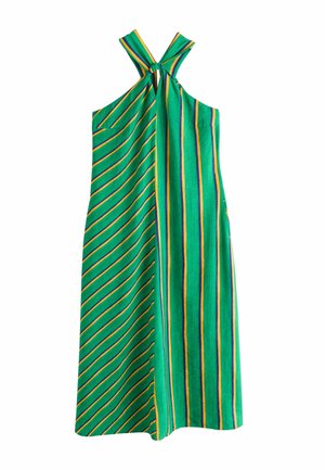 Kvinde i en grøn- og gulstribet halterneck-kjole står på en balkon med udsigt over en kystby under en klar blå himmel.