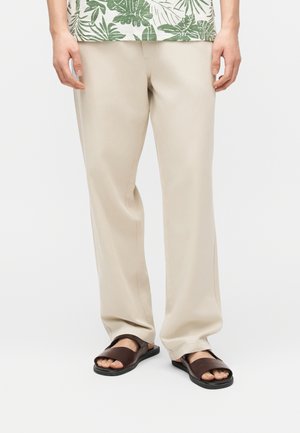 Jambes portant un pantalon beige à jambes larges, des sandales marron à enfiler et une chemise à imprimé tropical vert et blanc visible à la taille.