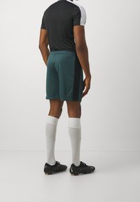 Hombre con camiseta deportiva en blanco y negro, pantalones cortos color teal con una franja negra, calcetas blancas hasta la rodilla y botas de fútbol negras, de pie de espaldas.