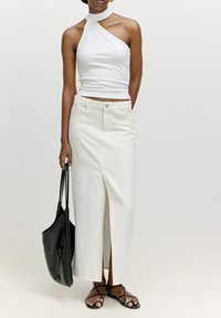 Witte haltertop in combinatie met een witte denimrok met een split aan de voorkant. Accessoires omvatten een zwarte tote bag en zwarte sandalen met banden.
