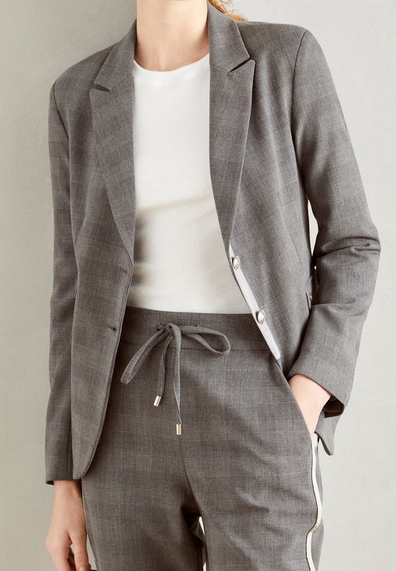 Femme portant un blazer à carreaux gris ajusté et un pantalon à cordon assorti avec des bandes blanches sur les côtés, associé à un top blanc ajusté.