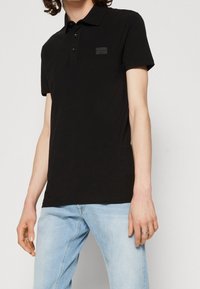 Černé polo tričko s krátkým rukávem vyrobené z hladké tkaniny, s položením na tři knoflíky a malou texturovanou nášivkou na hrudi.