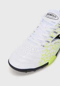 Fehér és neon sárga sportcipő fűzőkkel, perforált orr résszel, fekete talppal, és "Joma Maxima" logóval a nyelvén.