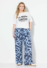 Vit t-shirt med svart text, kombinerad med vida blå leopardmönstrade byxor. Materialet verkar mjukt; avslappnad stil. Accenterad med ljusa skor.