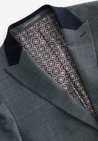 Blazer avec un tissu extérieur à carreaux bleus et des accents marron, complété par un col en côtes bleu marine. L'intérieur présente un motif géométrique en bordeaux et crème.