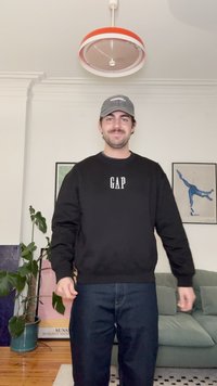 Fekete pulóver fehér "GAP" logóval, sötétkék farmernadrággal párosítva. A szobában növények és absztrakt falikép található.