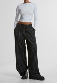 Gekürztes weißes Langarmoberteil und weit geschnittene schwarze gestreifte Hose mit vorderen Falten. Schwarze klobige Stiefel setzen einen Kontrast zum Outfit.